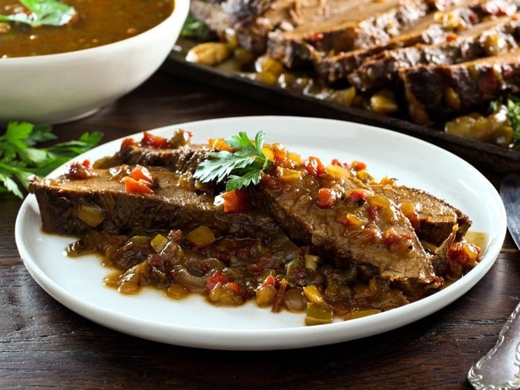 Two slices of West African Brisket on sauce topped with herbs on plate with bowl of sauce, brisket in background.