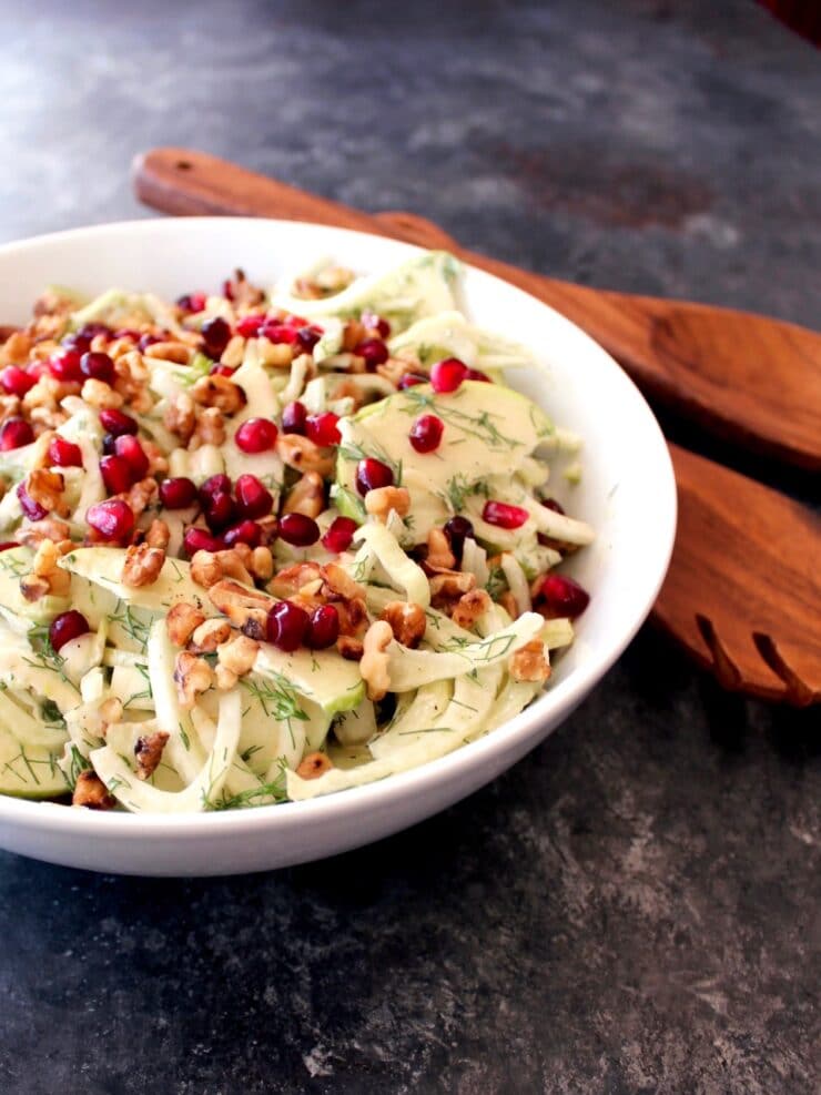 Close up of a bowl of Fennel Apple Salad with Cider Tahini Dressing, topped with pomegranate seeds, with wooden utensils.