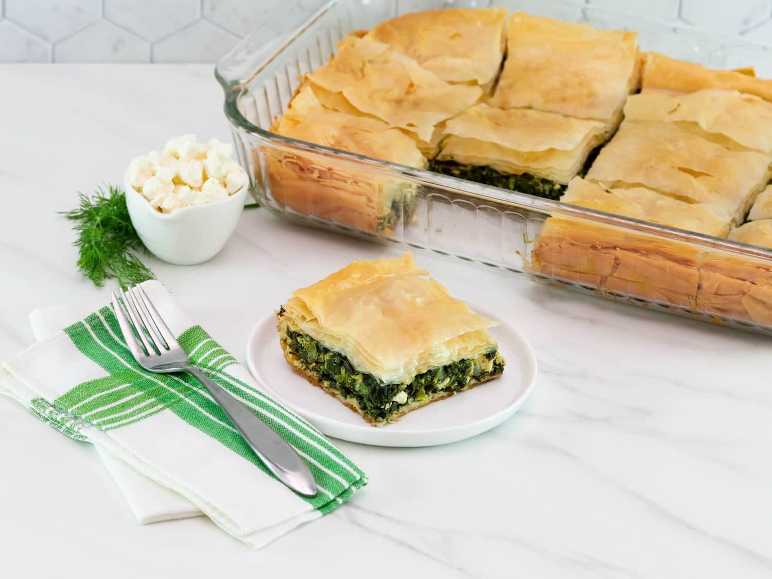 Horizontal shot of a slice of spanakopita on a white plate, a baking dish filled with the remaining spanakopita is sitting in the background.