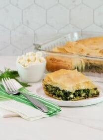 Vertical shot of a slice of spanakopita on a white plate, a baking dish filled with the remaining spanakopita is sitting in the background.