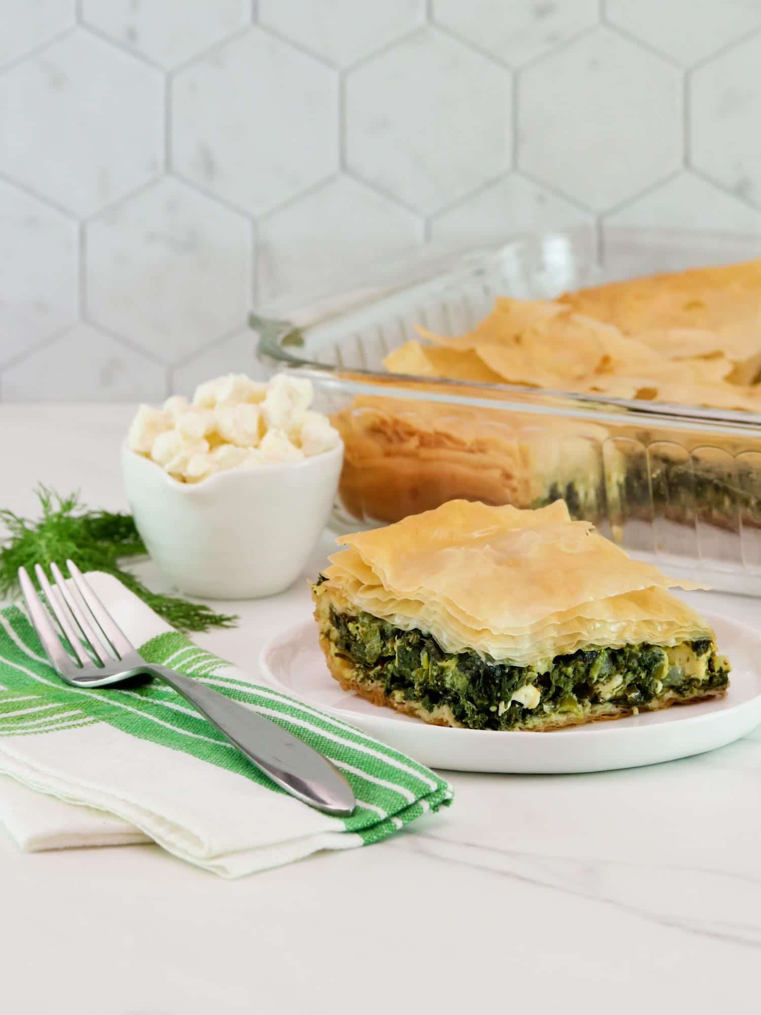 Vertical shot of a slice of spanakopita on a white plate, a baking dish filled with the remaining spanakopita is sitting in the background.