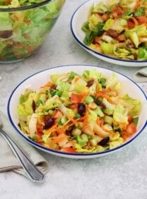 Horizontal shot of a colorful salad with fork and cloth napkin, second dish of salad and large glass salad bowl with towel in background, on a grey marble surface.