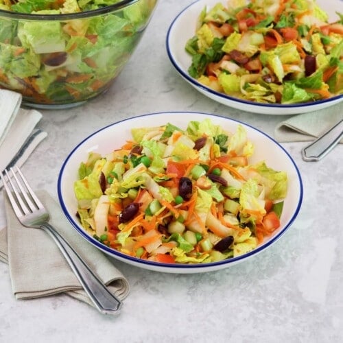 Horizontal shot of a colorful salad with fork and cloth napkin, second dish of salad and large glass salad bowl with towel in background, on a grey marble surface.