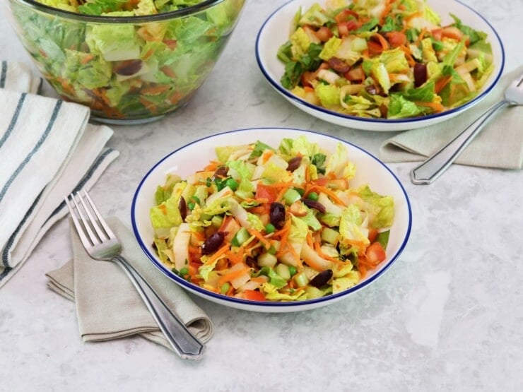 Horizontal shot of a colorful salad with fork and cloth napkin, second dish of salad and large glass salad bowl with towel in background, on a grey marble surface.