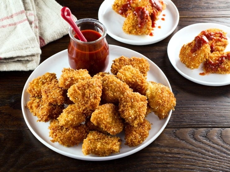 Plate of schnitzel nuggets with red sriracha sauce on wooden table, towel in background.