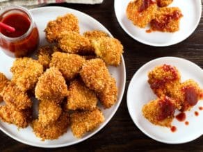 Overhead horizontal shot of smoky panko schnitzel bites with small glass carafe of red sriracha sauce on white plate.