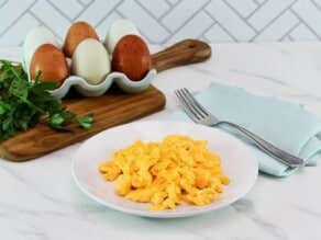 Horizontal shot of a plate of perfect scrambled eggs - moist, fluffy, bright yellow - on a white plate. Linen napkin in background with wooden cutting board, brown and white eggs in a carton, and fresh parsley.