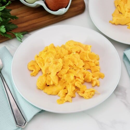 Fluffy scrambled eggs on a plate with fork and cloth napkin, on a marble countertop. Wooden cutting board with egg carton - brown and white eggs, and fresh parsley. Another plate of scrambled eggs in background.