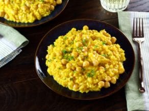 Front angled shot of creamy Saffron Chickpea Risotto on dark blue plate, another plate of risotto, fork, cloth napkin and short decorative wine glass in background.