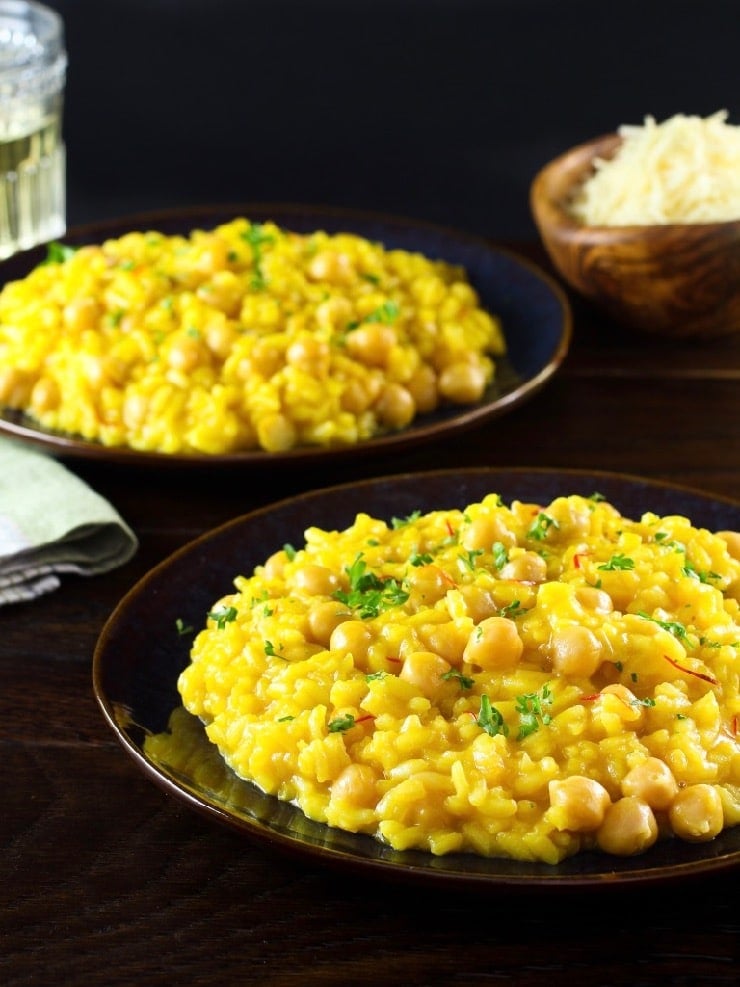 Vertical long shot of creamy Saffron Chickpea Risotto on dark blue plate, another plate of risotto, fork, cloth napkin and short decorative wine glass in background. Wooden bowl of grated parmesan on table behind.