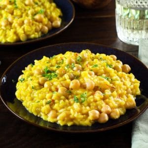 Front angled shot of creamy Saffron Chickpea Risotto on dark blue plate, another plate of risotto, fork, cloth napkin and short decorative wine glass in background.