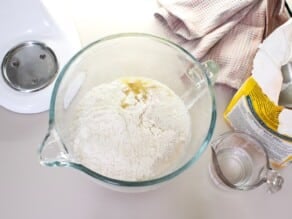 Flour, oil, and other ingredients in a glass mixing bowl ready for the mixer. Small measuring cup, bag of flour and linen towel on the side.