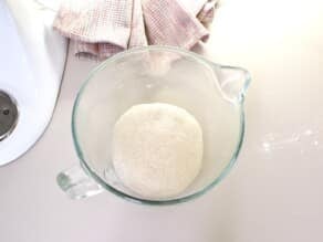 Ball of dough in a stand mixer glass bowl on countertop with linen napkin in background.