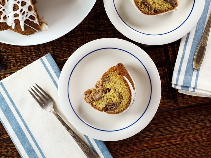 Overhead shot - slice of sour cream coffee bundt cake with drizzled white icing on plate with fork, napkin beside it on wooden table.