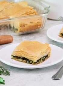 Horizontal shot of a two white plates containing square slices of vegan spanakopita. A glass dish with the remaining spanakopita is in the background to the left.