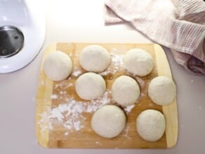 Eight dough rounds on a wooden cutting board surface sprinkled lightly with flour. Linen towel in background.