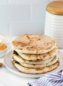 Horizontal close up - Pile of freshly baked pita breads on a plate, striped blue and white linen napkin in foreground, two small dishes of dip in background - hummus and yogurt dip - with a wooden spoon and a flour canister in the background, as well.