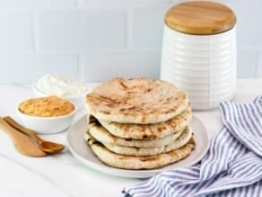 Horizontal close up - Pile of freshly baked pita breads on a plate, striped blue and white linen napkin in foreground, two small dishes of dip in background - hummus and yogurt dip - with a wooden spoon and a flour canister in the background, as well.