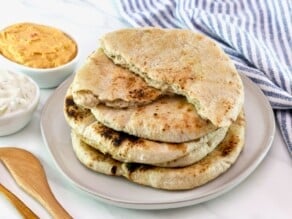 Horizontal image - stack of sourdough pita breads on a plate, top two pits are ripped in half. Striped towel and two dishes of dip in background - one hummus, one yogurt dip. Wooden spoon in foreground. All on a marble countertop.