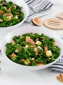 Overhead tilted horizontal shot of two white bowls filled with fig salad - fresh fig halves tossed with dark green curly kale, fried halloumi cheese, and walnuts. Linen napkins and fork, two wooden spoons for serving decorate the white marble countertop, alongside fresh fig halves scattered beside the bowls.