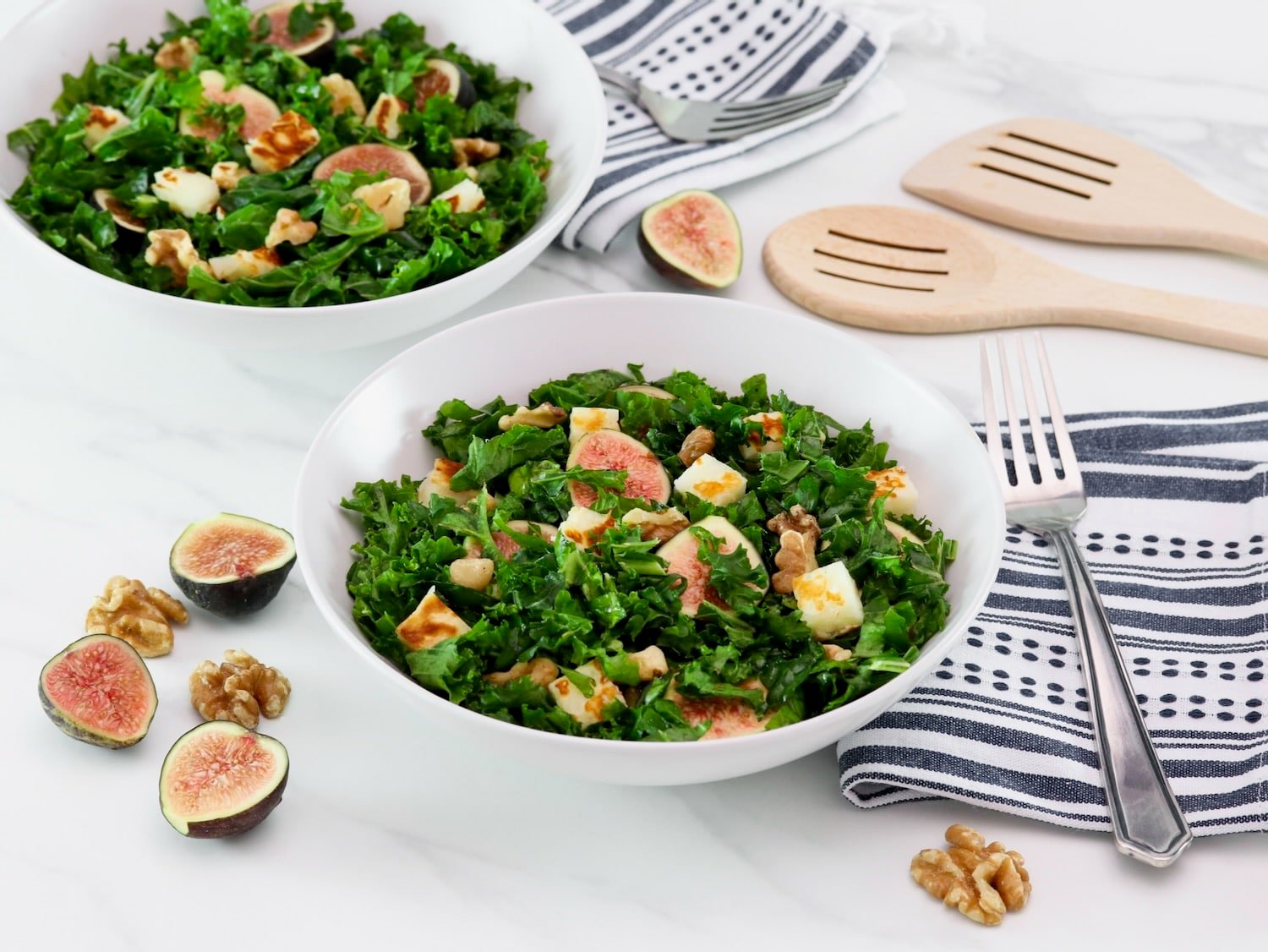 Overhead tilted horizontal shot of two white bowls filled with fig salad - fresh fig halves tossed with dark green curly kale, fried halloumi cheese, and walnuts. Linen napkins and fork, two wooden spoons for serving decorate the white marble countertop, alongside fresh fig halves scattered beside the bowls.