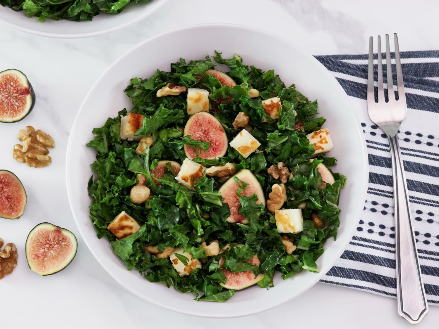 Overhead shot - large white bowl filled with fig salad with dark green curly kale, pan-seared halloumi cheese, and walnuts. Fork and linen napkin beside the bowl, fresh fig halves scattered across the marble countertop.