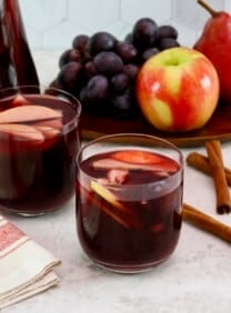 Close up shot - two unstemmed wine glasses filled with red wine Thanksgiving sangria, fresh fruit slices - apples and grapes floating in the cups. Linen towel beside the cups. On a white marble countertop with three cinnamon sticks, a platter of fruit - apple, grapes, pears - and a large pitcher of sangria in the background.