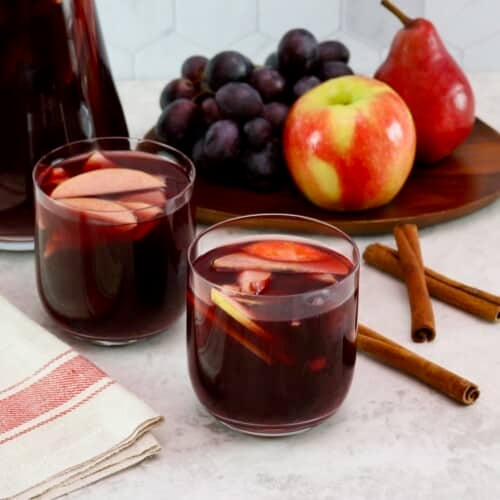 Close up shot - two unstemmed wine glasses filled with red wine Thanksgiving sangria, fresh fruit slices - apples and grapes floating in the cups. Linen towel beside the cups. On a white marble countertop with three cinnamon sticks, a platter of fruit - apple, grapes, pears - and a large pitcher of sangria in the background.