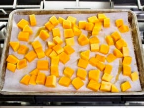 Overhead shot - tray of butternut squash cubes drizzled with maple syrup on a parchment-lined baking sheet, resting on a stovetop.