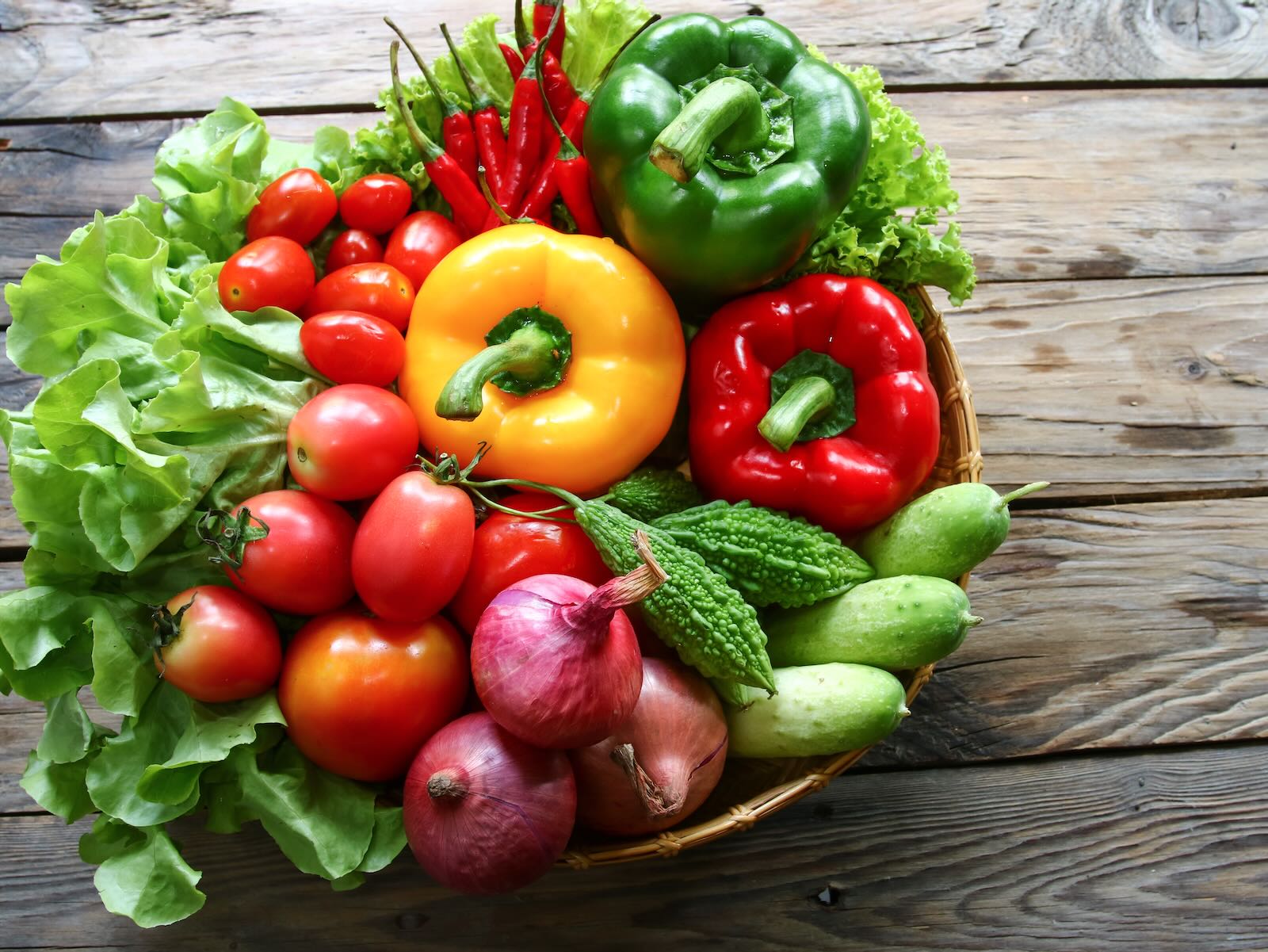 Horizontal image of a bowl, sitting on a wooden table, filled with an assortment of fresh, colorful vegetables.