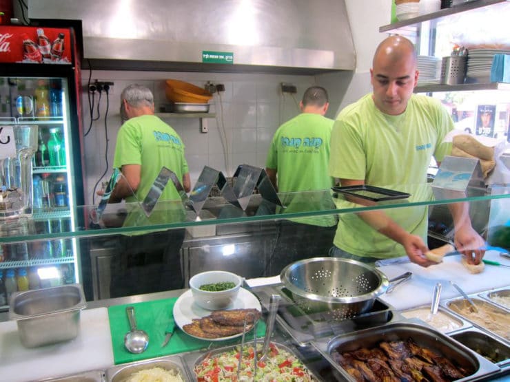 Mana Mana falafel restaurant - man is making a pita sandwich at counter with salad fixins. Two workers are in the kitchen behind him.