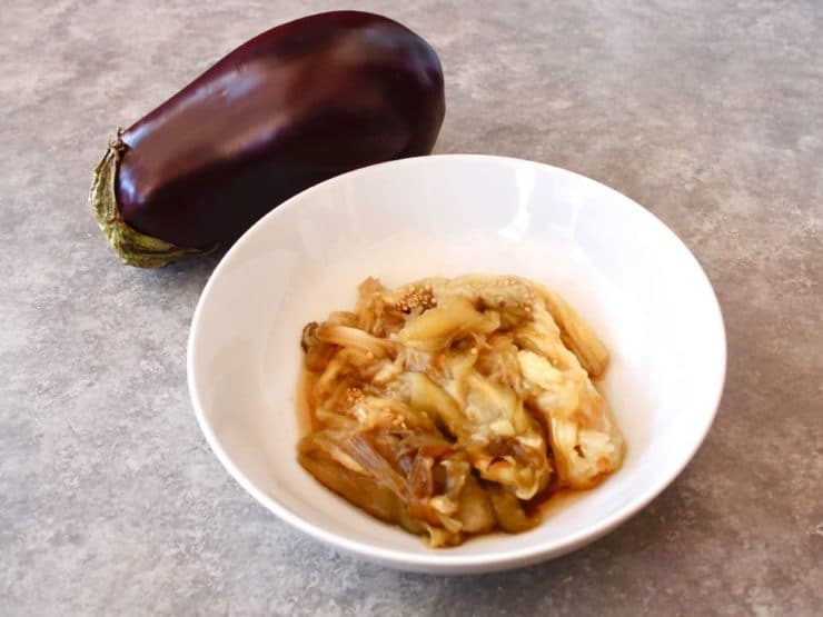 A whole eggplant beside a bowl of cooked eggplant on a grey countertop