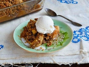 Horizontal cut image of delicious Oat Nut Apple Crisp dessert with a golden brown crumble and ice-cream topping served in a white ceramic dish