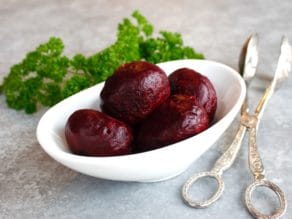 Roasted peeled beets in a white bowl with fresh parsley and antique silver tongs.