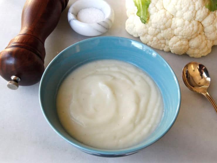 Image of a Cauliflower Soup on a bowl with a spoon, pepper crusher, salt, and cauliflower in the side