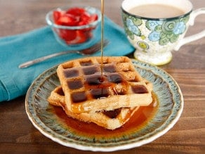 Close-up of two Tahini Waffles drizzled and soaked with syrup and a cup of coffee and fork in the background