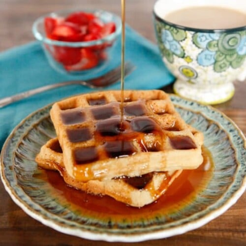 Close-up of two Tahini Waffles drizzled and soaked with syrup and a cup of coffee and fork in the background