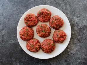 7 burger patties on a white plate with grey concrete background.