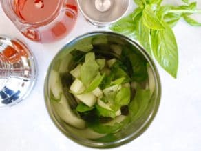 Cucumber and basil in cocktail shaker with basil, jigger, ginger agave syrup carafe, and shaker lid surrounding on white cutting board.