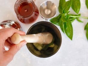 Muddling cucumber and basil in cocktail shaker with fresh basil, jigger, ginger agave syrup carafe, and shaker lid surrounding on white cutting board.