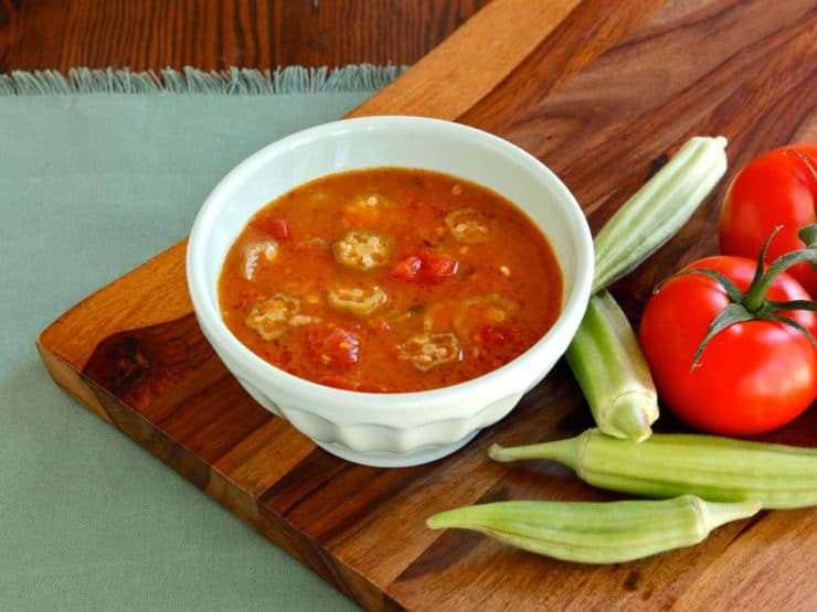 A horizontal shot of Okra soup served on a white bowl with fresh okra and tomato on side of the bowl