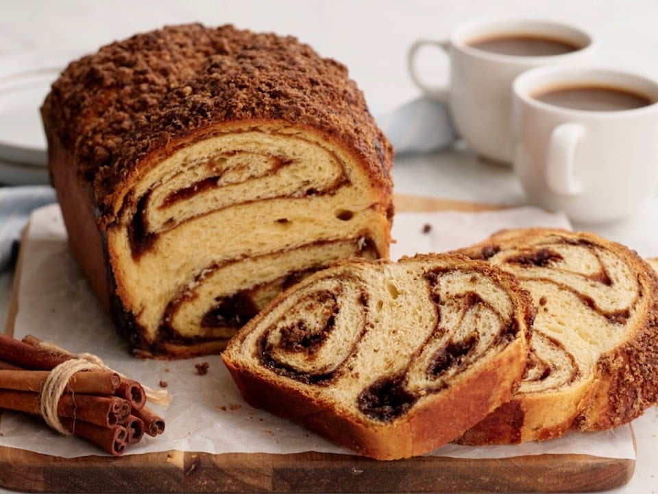 Horizontal image - Cinnamon Babka sliced on cutting board with cinnamon stick bundle. coffee and towel in background.