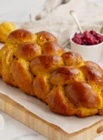 Horizontal image of pumpkin challah bread loaf on wooden cutting board, surrounded by fruits in each corner.