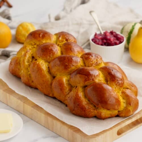 Horizontal image of pumpkin challah bread loaf on wooden cutting board, surrounded by fruits in each corner.