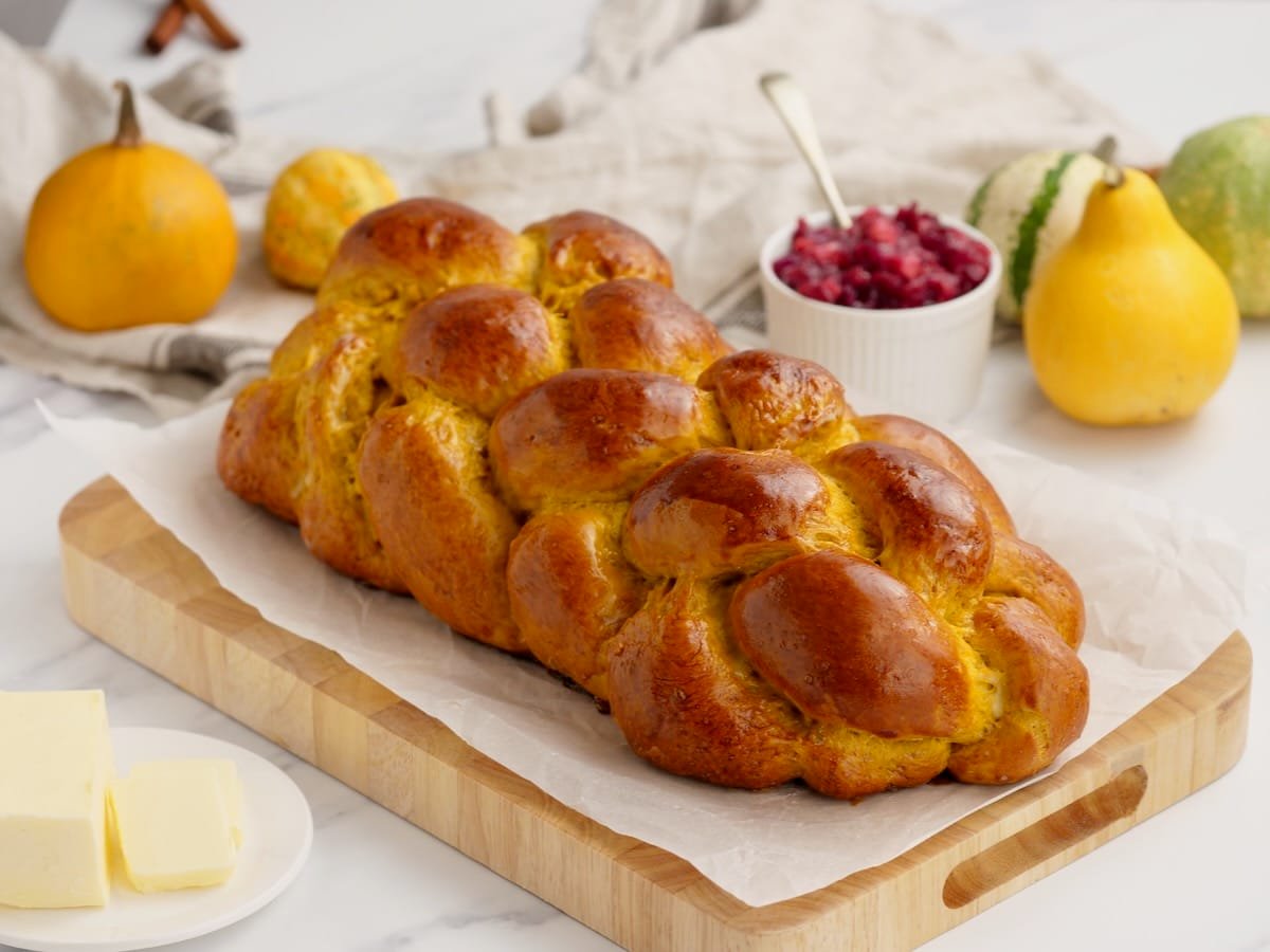 Horizontal image of pumpkin challah bread loaf on wooden cutting board, surrounded by fruits in each corner.