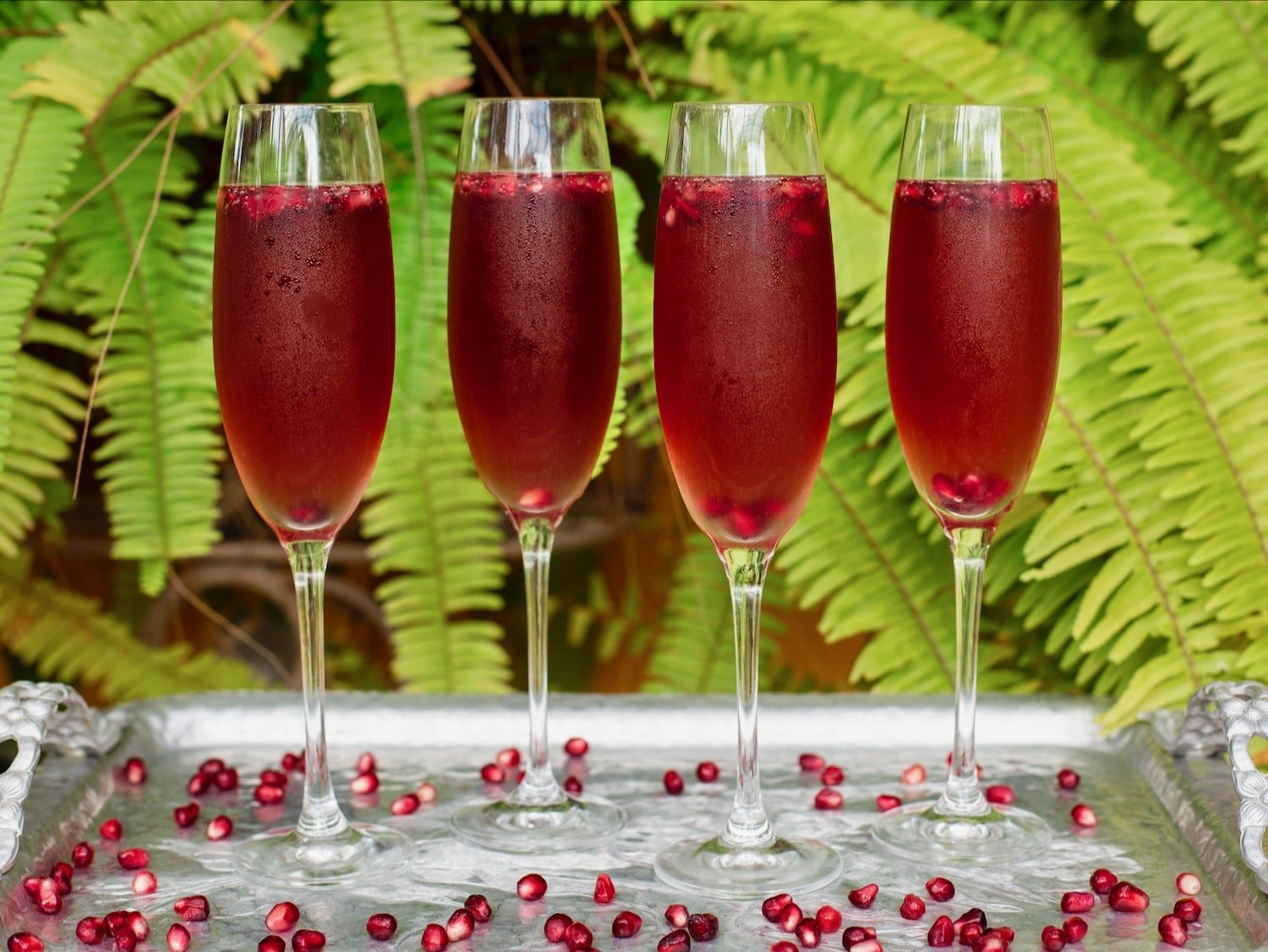 Vertical shot of four champagne flutes filled with a bright red pomegranate cocktail. Pomegranate arils are floating in the cocktails and scattered around the table.