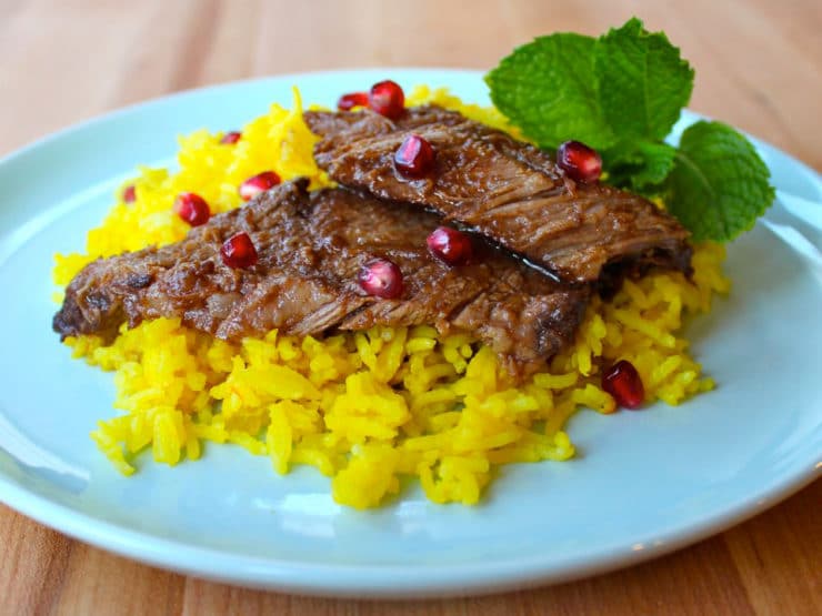 Slices of Pomegranate Molasses Brisket on top of saffron rice with pomegranate arils and mint garnish on blue plate atop wooden cutting board.