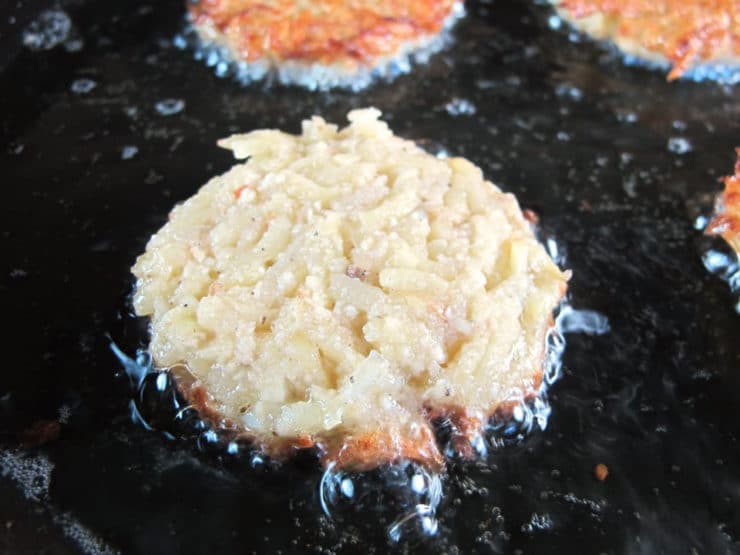 Shredded latke half-fried turning golden in sizzling oil in black skillet.