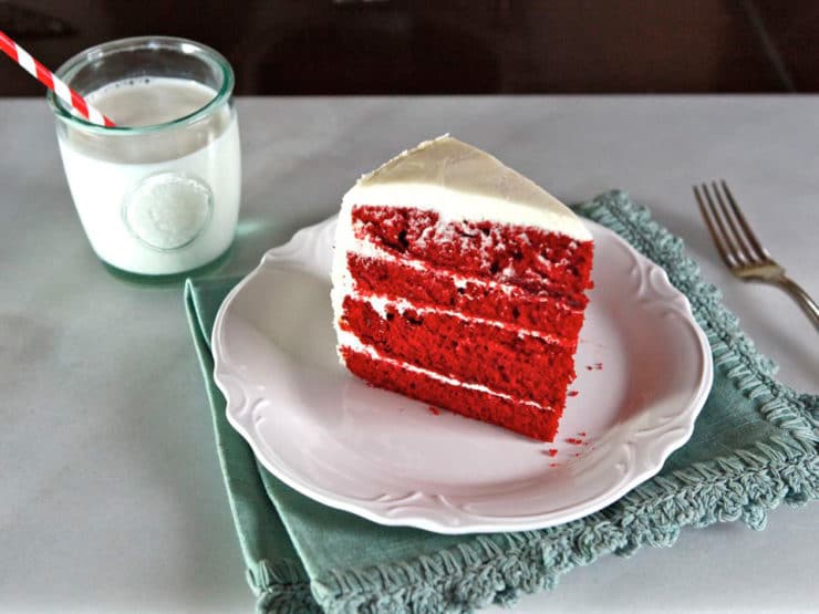 A slice of moist and decadent red velvet cake with cream frosting with a milk drink on the side