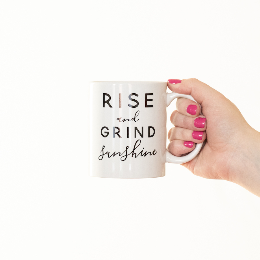 White coffee mug with RISE and GRIND sunshine printed on it in black bold and cursive letters